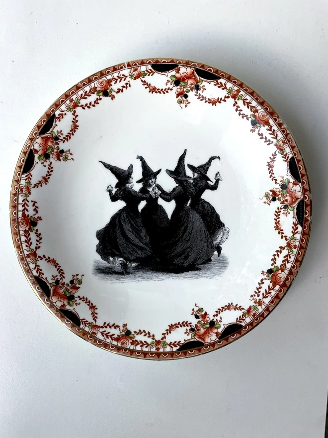 a coven of 4 witches dance hand in hand, the image is victorian in black and white and is printed directly onto an ornate vintage plate