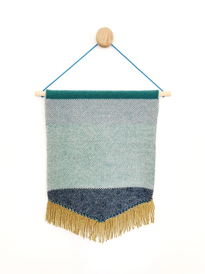 A product image showing the reverse of a jade green  knitted wall hanging. The back of the banner is knitted into a navy and white/ green and white birdseye check and is edged with a gold bullion trim. The whole flag is threaded onto a wooden dowelling and  suspended from a wooden wall dot by a bright teal nylon cord.