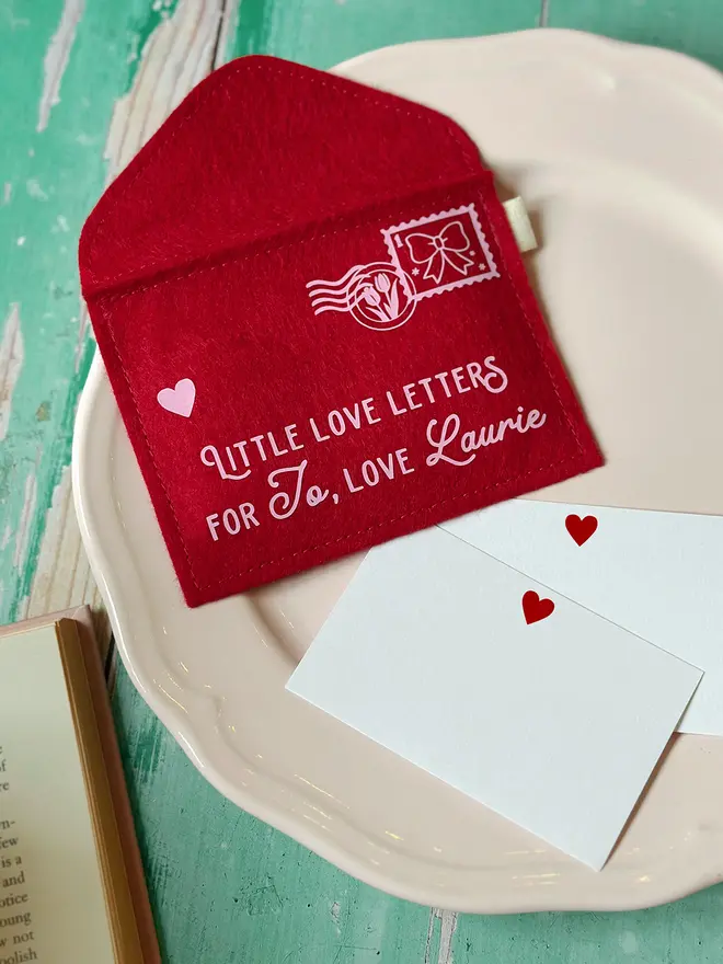 handmade personalised red felt love letters envelope displayed on a vintage-style plate with small heart cards, beside an open book, styled on a rustic green wooden table for Valentine’s Day.