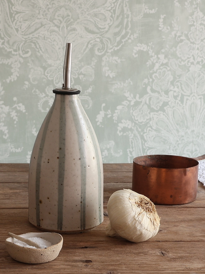 
Handmade Ceramic Stoneware Oil Pourer in Blue and White Stripe sat on a wooden surface next to a bulb of garlic, a small handmade ceramic dish containing salt and a brass measuring cup. 
