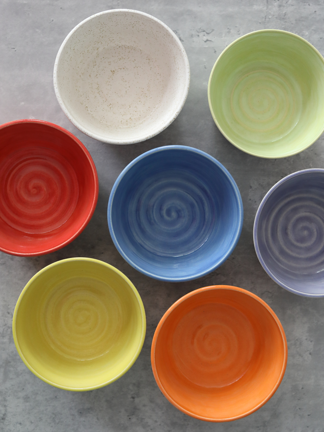 Seven colourful glazed ceramic bowls arranged on a grey surface, viewed from above at an angle, in yellow, red, orange, green, blue, purple, and speckled white, each featuring a swirl pattern on the interior.