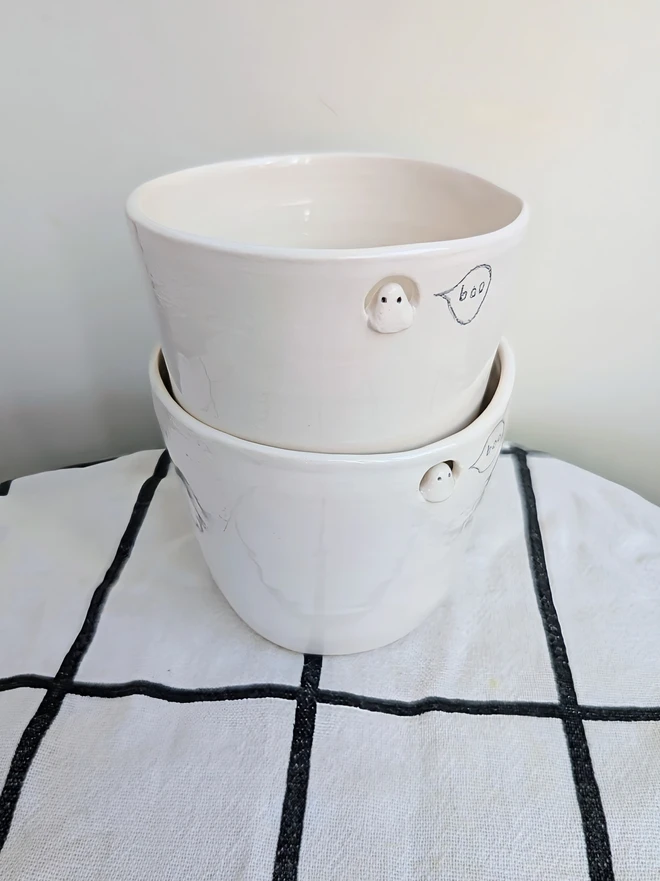 stacked small ghost cups with finger dents in white handmade ceramic with a tiny sculpted ghost in a cut out near the top of the beakers with a hand painted bubble with the word boo in