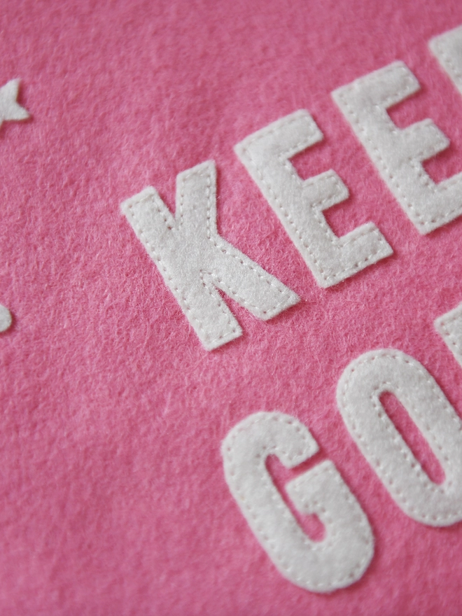 A pink felt banner flag lays on white fabric. White felt stars are scattered around the words Keep Going in the centre of the flag.