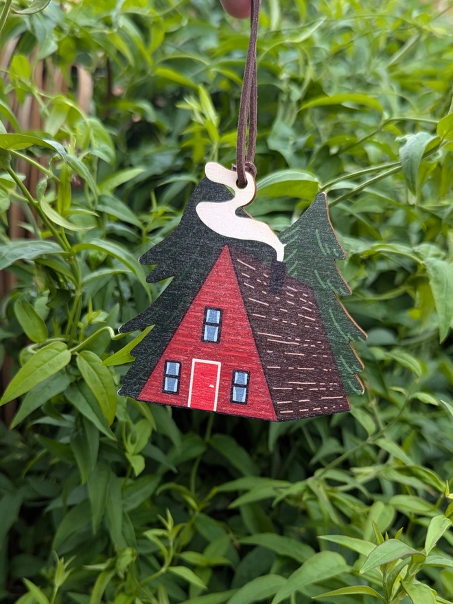 Red cabin wooden Christmas decoration, with smoke billowing from the roof and 2 pine trees.