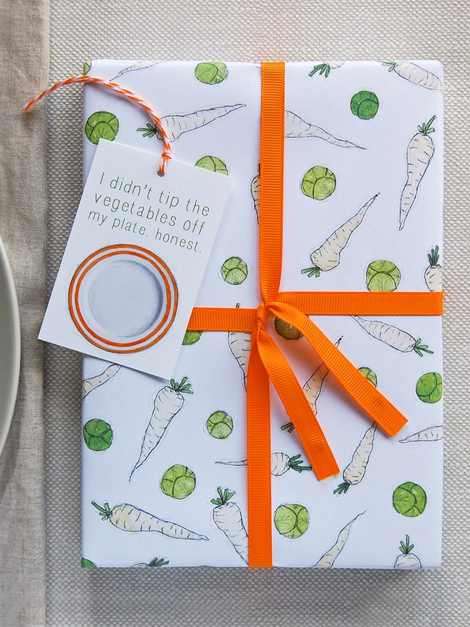 A Christmas gift wrapped in fun illustrated sprouts and parsnips wrapping paper is tied with orange ribbon.