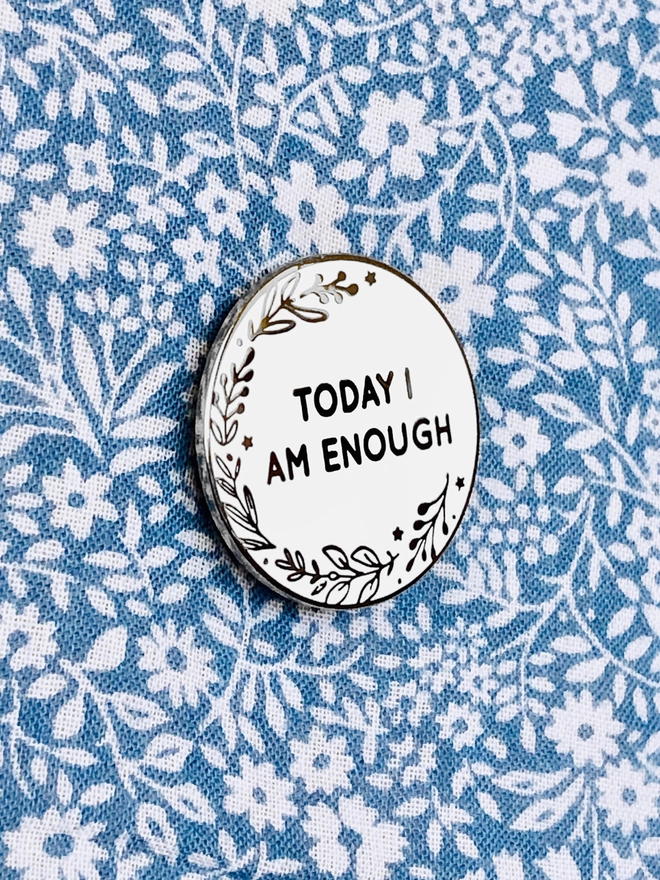 A round white enamel pin with a botanical design and the words "Today I Am Enough" is pinned to blue floral fabric.