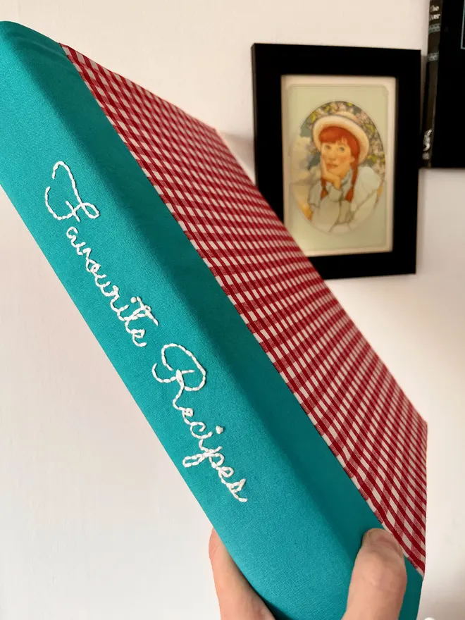 Hand holding up a red gingham-covered ring binder with personalisation embroidered on the teal spine