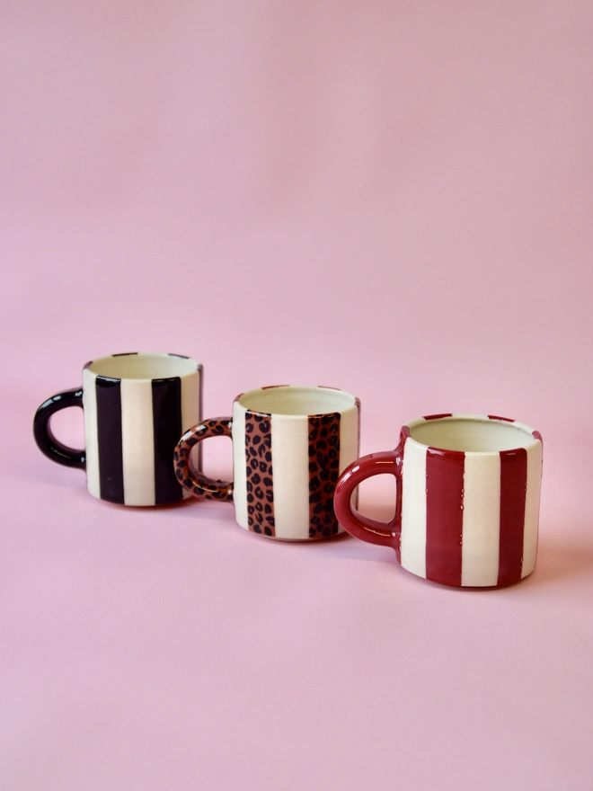 Set of three handmade ceramic mugs with bold striped designs in black, red, and leopard print, arranged on a soft pink background.