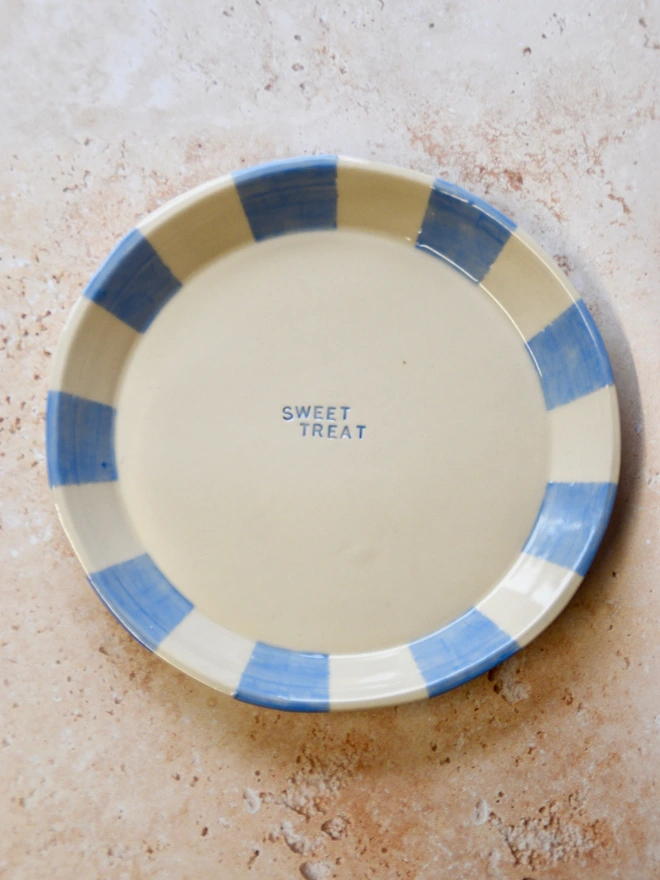 Handmade ceramic plate with blue striped rim and “Sweet Treat” text in the centre, photographed on a neutral stone background.