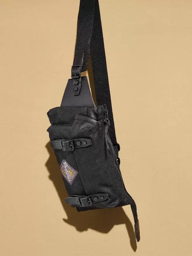 Black sling bag with black leather trim hanging against taupe backround