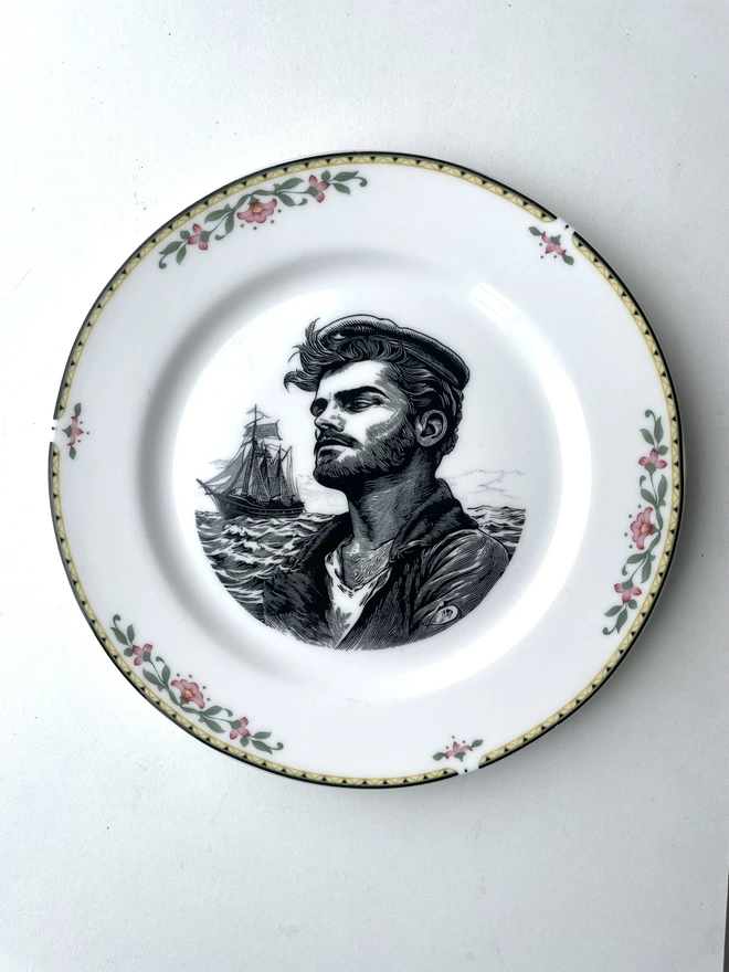 vintage plate with an ornate border, with a printed vintage illustration of a sailor man in the middle with the ocean and his ship behind him