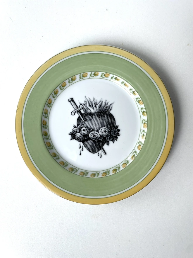 a vintage plate with a white and gold ornate border, the centre is lemon yellow and printed on that is a black illustration of a sacred heart