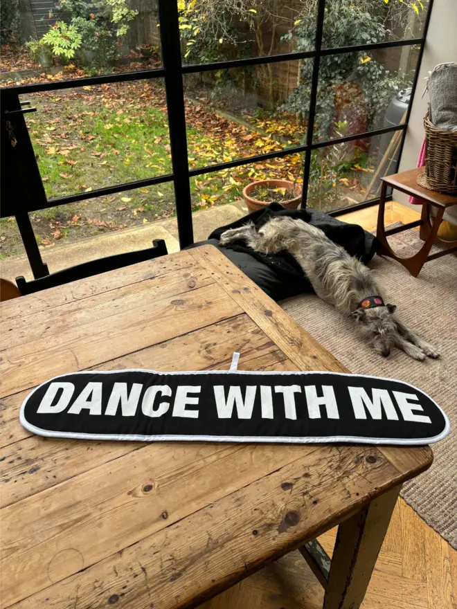 Black oven glove that says DANCE WITH ME on it laying on a wooden table with dog asleep on floor in front a large window in the background