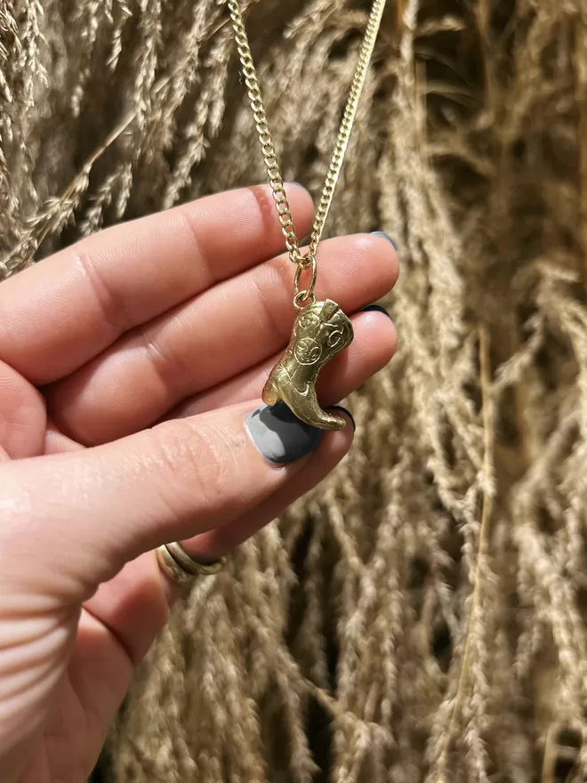 Image of hand carved cowboy boot pendant hanging on a chain, being held by a female hand wearing grey nail polish. The background of the image is dried pampas grasses. 