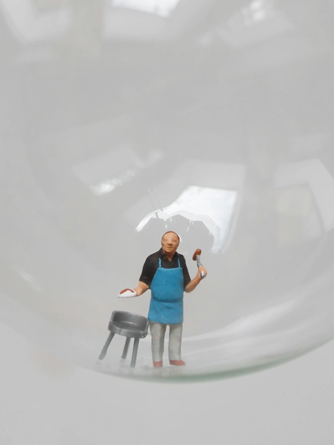 Miniature figure of a man at a bbq in a glass Christmas bauble - close up 