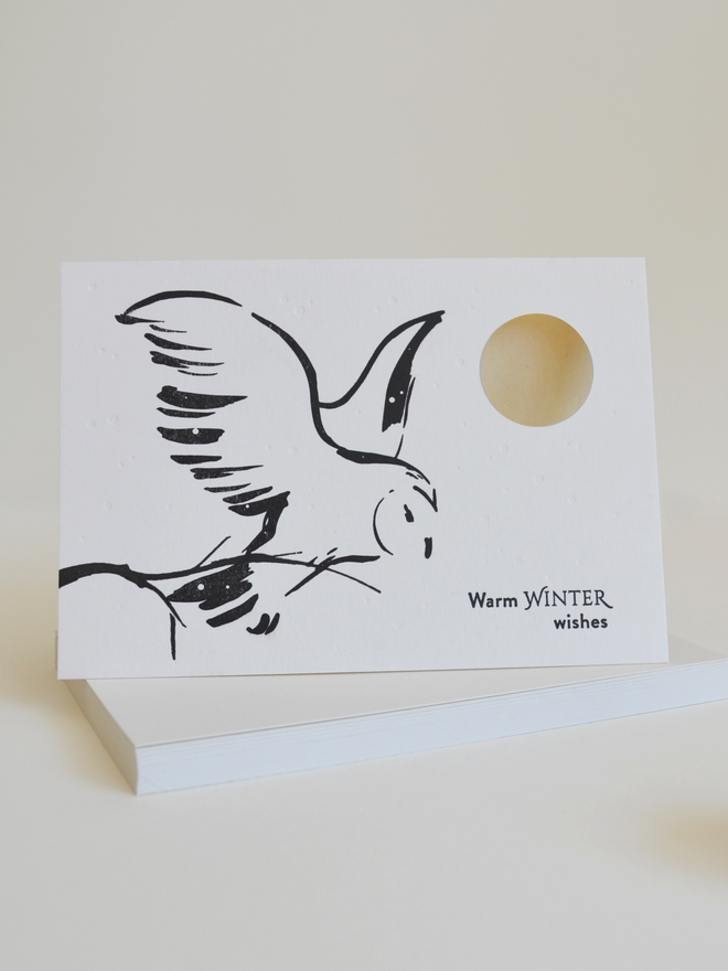 black and white winter-themed greeting card with a snowy owl in flight against a cut-out moon.