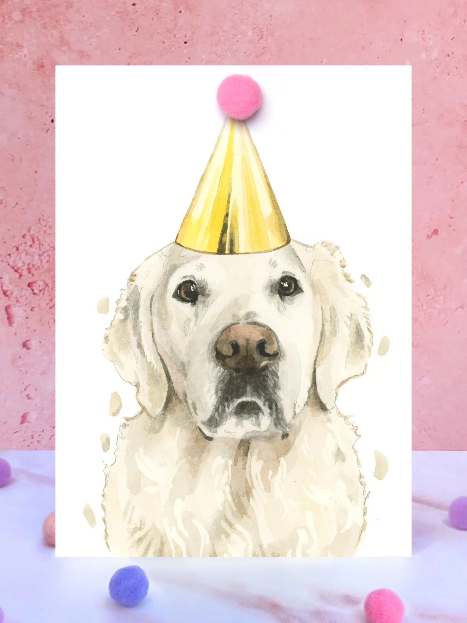 A greeting card featuring a hand painted design of a cream golden retriever, stood upright on a marble surface surrounded by pompoms. 