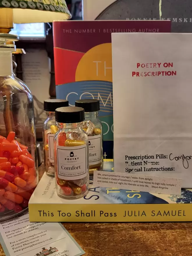 Glass bottles of comfort poetry pills in yellow, blue and orange,and a "poetry on prescription" bag surrounded by books, pills, and quotes on slips of paper