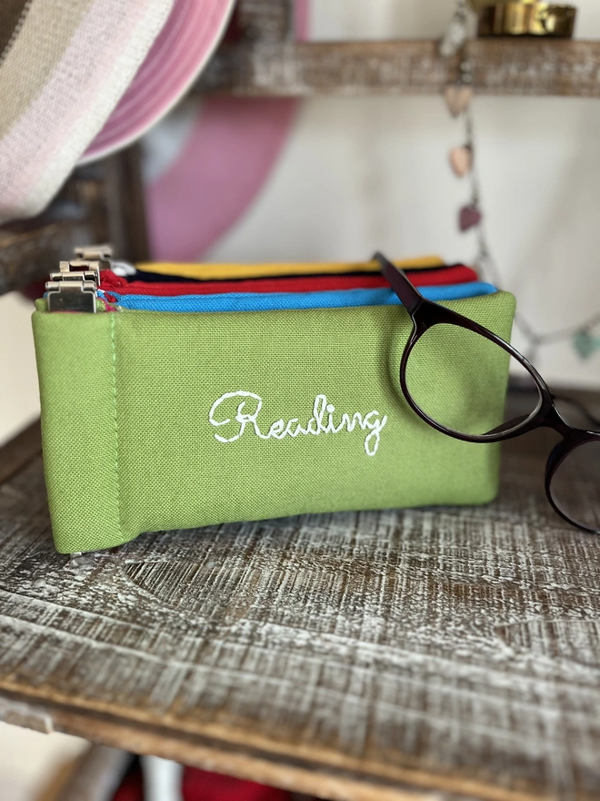 Bright green soft cotton glass pouch with spectacles hanging over it. Four more cases in alternative bright colours stacked behind it. 