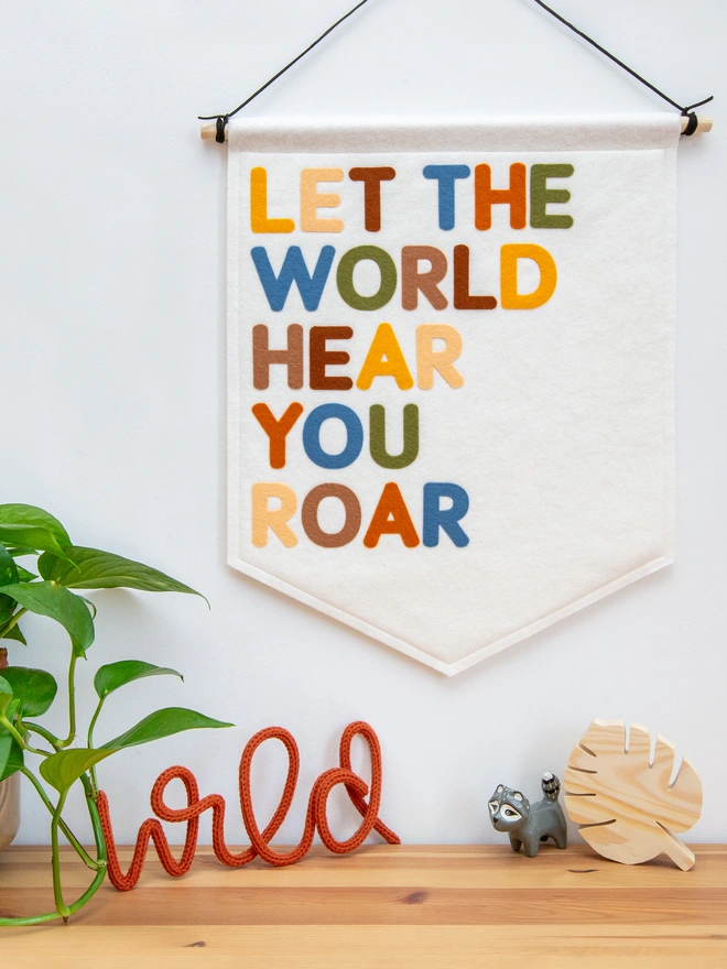 White felt wall banner with the words "LET THE WORLD HEAR YOU ROAR" written on it in a mix of earthy, jungle themed colours.