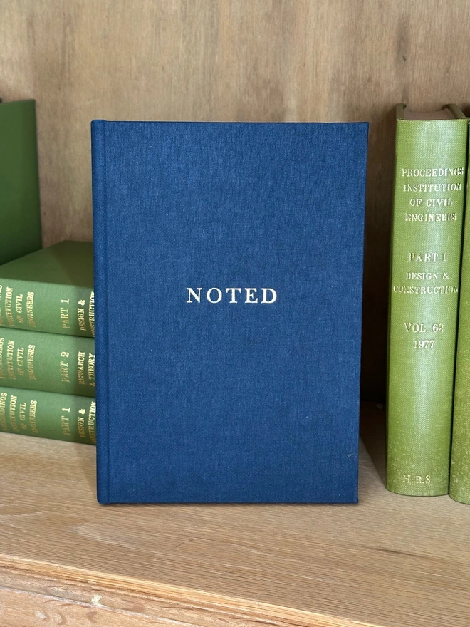 A navy linen notebook printed with 'Noted' in gold lettering rests on a wooden bookshelf next to numerous green books.