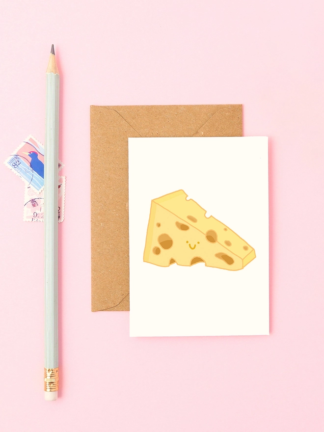 Mini cheese card with smiling Swiss cheese illustration, shown on a pink background with a kraft envelope and pale green pencil.