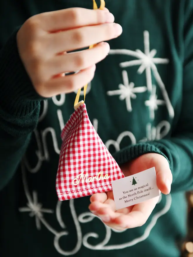 A child wearing a green Christmas jumper holds a red gingham personalised fabric fortune cookie with the name “Martha” in gold, revealing a tiny wish card inside that reads “You are as magical as the North Pole itself… Merry Christmas!”