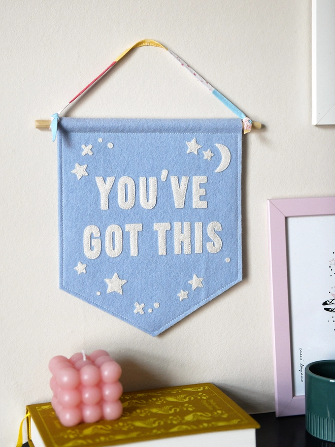 A blue felt wall hanging, with the words You’ve Got This and white felt stars, hangs on a white wall from its ribbon hanger.