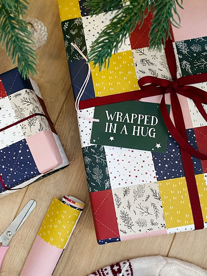 Gifts wrapped in a traditional patchwork design wrapping paper are on a wooden floor beneath a Christmas Tree.