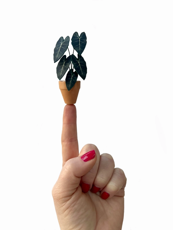 A miniature replica Alocasia Frydek ornament made from paper in a terracotta pot balanced on a finger