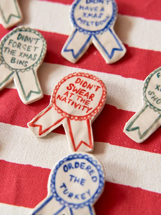 A selection of Red, Green & Blue Handmade Ceramic Christmas Award Badges, handpainted with different phrases, atop a striped red and white tablecloth