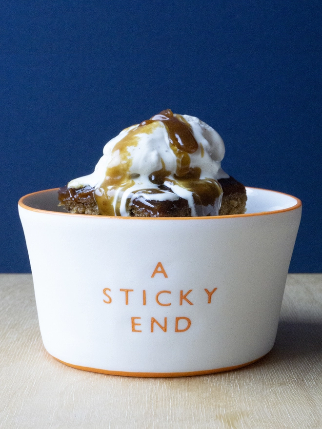 Ceramic porcelain pudding bowl with A Sticky End written on the side. A white pudding bowl with orange rim containing sticky toffee pudding.