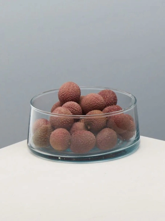 Low Glass Dish, a small glass dish sitting on a white counter top against a baby blue backdrop. It is filled by lychees. 
