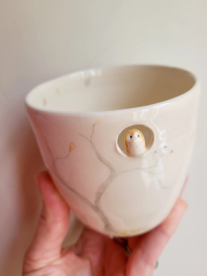 ivory cream ceramic handleless cup with a tiny ceramic owl in a cut out hole near the rim there are hand painted trees and orange and yellow leaves 