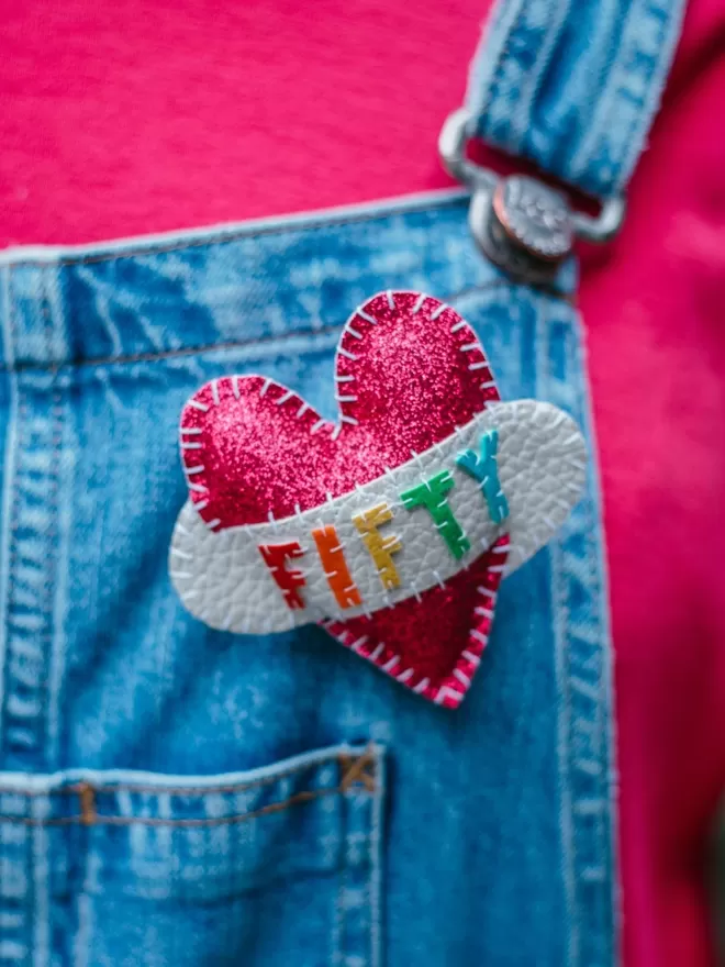 Pink glittery heart shaped brooch with FORTY spelt out in rainbow coloured lettering on a white scroll, pinned to blue denim dungarees
