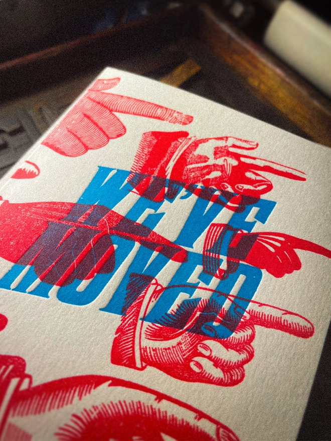 A double-sided, hand-printed letterpress artisan moving house announcement card with bold blue “We've Moved” typography and fluorescent red pointing hand illustrations (fists) on thick premium white card.