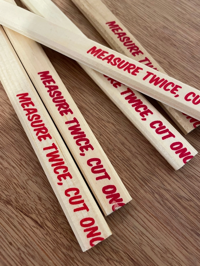 Wooden flat carpenters pencils laid on a wooden table. The pencils have a slogan Measure Twice Cut Once screenprinted at one end in red ink, but the joke is that last word has been cut off, or mispositioned. 