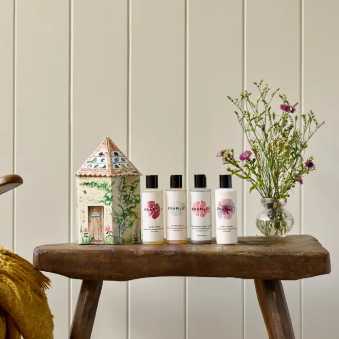 card board box in the design of an illustrated bird house, next to 4 boxes of product with white labels and black lids, and a glass vase with green and purple flowers, all sitting on a wooden stool