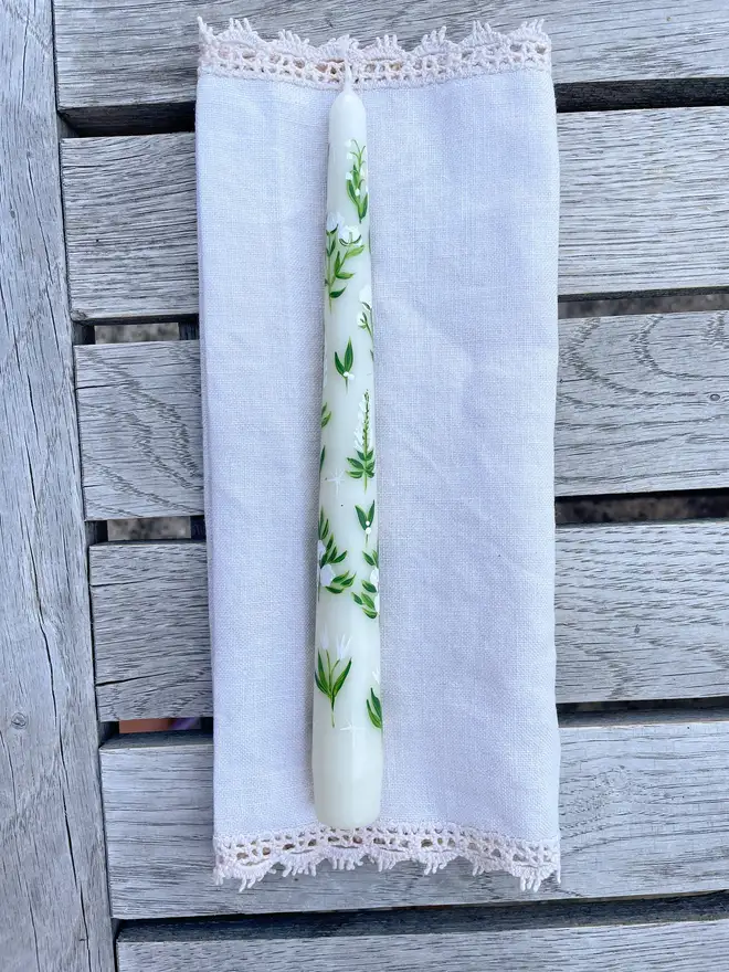 White Florals Dinner Candle, a white candle on a napkin on a wooden surface. It's green floral design wraps around it. 