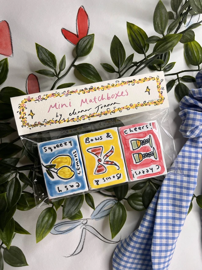 A collection of three hand-painted matchboxes with playful and colorful designs: one features two glasses with the word "Cheers!" written in bold lettering, another displays bright yellow lemons with "Easy Squeezy" written alongside, and the third showcases a red bow with the phrase "Bows & Arrows." Each matchbox is crafted with vibrant illustrations and creative lettering. By Eleanor Joanna