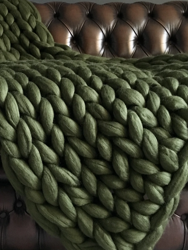 A close up of a willow green merino arm knitted blanket cascading down the side of a brown leather Chesterfield sofa