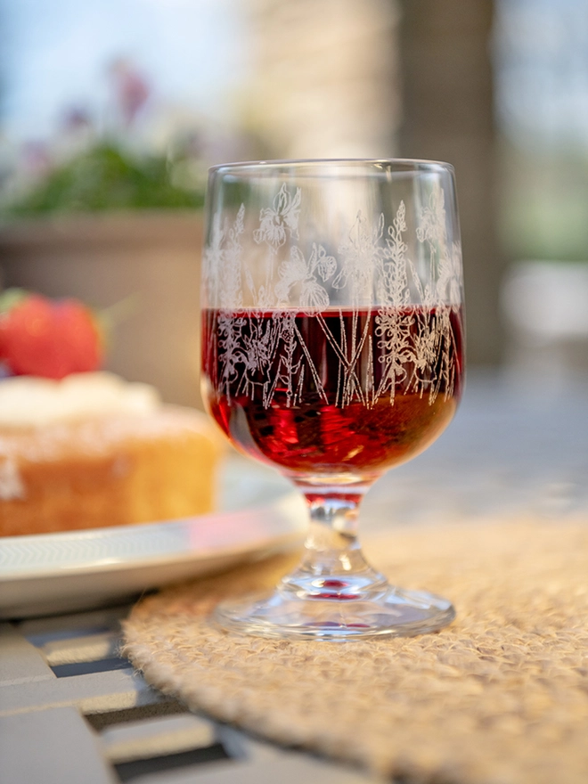 A short-stemmed bistro glass decorated with, illustrated poppies, hydrangeas, and sunflowers against a beige background.