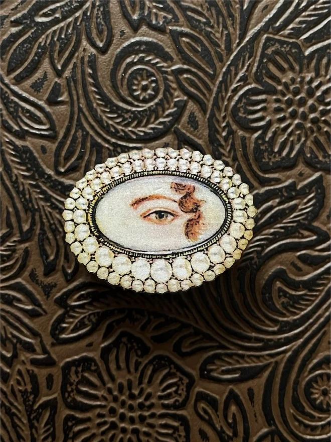 a lovers eye brooch, lovers eye is surrounded by pearls, this illustration is then printed onto wood and has a brass pin back