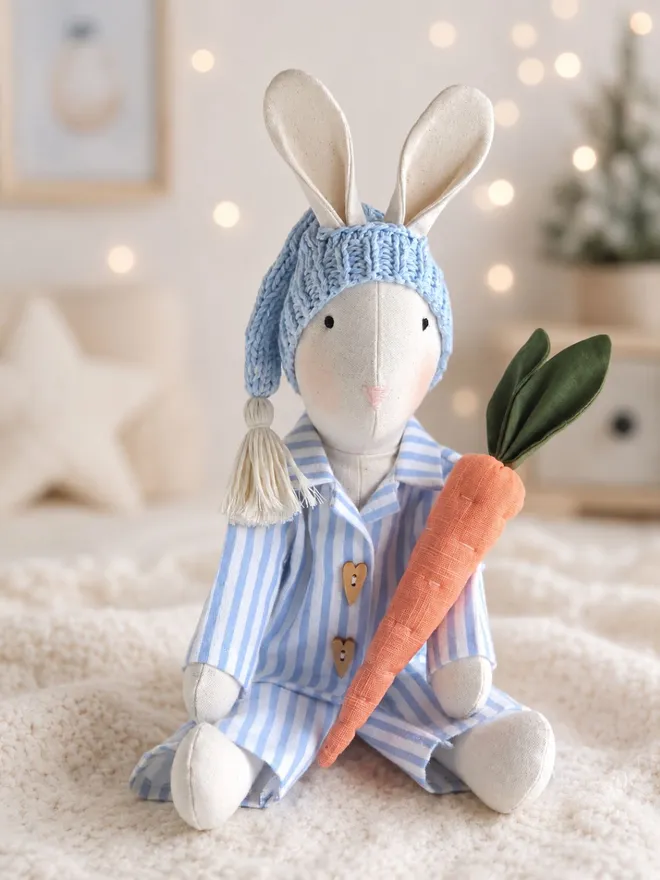 Handmade Gerald Bunny in blue striped Christmas pyjamas and knitted sleepy hat, holding a linen carrot in a softly lit festive nursery, hand crafted by Verity Isabelle.