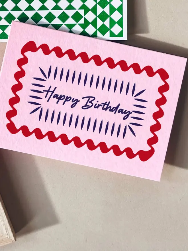 A pink hand-printed birthday card with a red wavy border and blue decorative lines around the handwritten words “Happy Birthday,” displayed on a table.