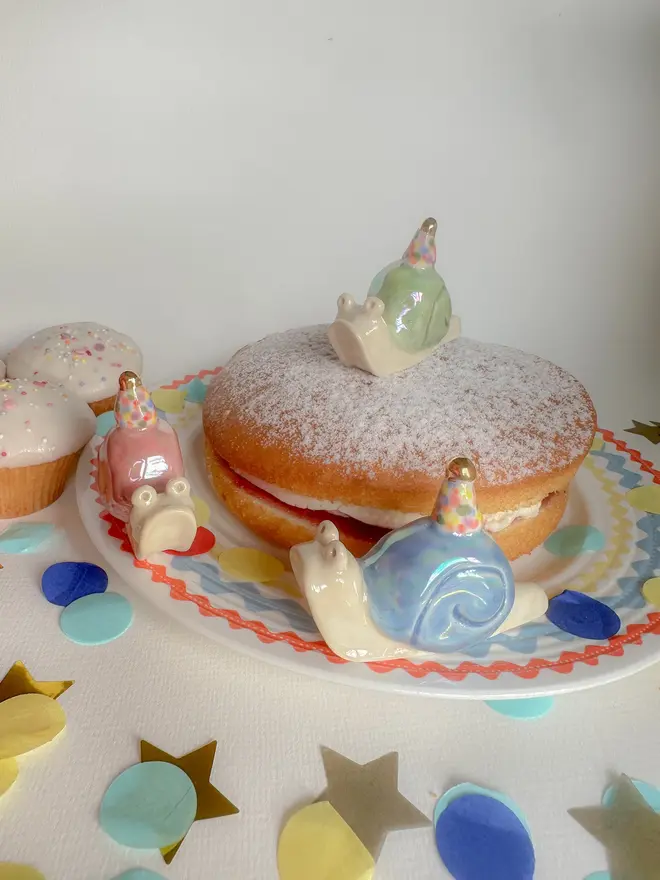 Handmade Porcelain Snail Cake Topper With Golden Hat, grouped together on a table with party decorations by a cake. 