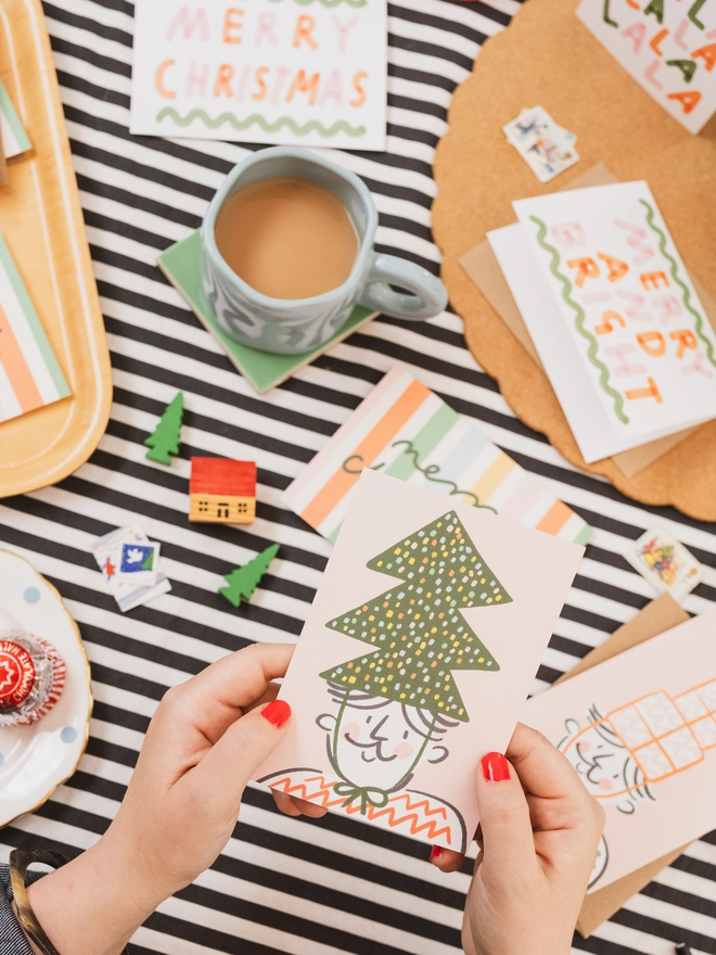 Image shows person holding a Christmas card which features an illustration of a man wearing a Christmas tree party hat.