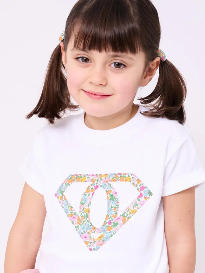 A girl wearing a white short sleeve t-shirt appliquéd with a superhero motif  featuring a letter O initial inside it. The appliqué is made from a floral Liberty print .