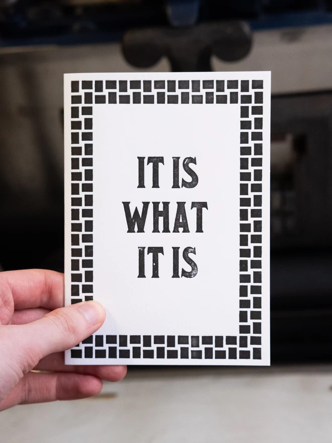 Letterpress card with text design "It is what it is," Surrounded by art deco style border of woven squares.