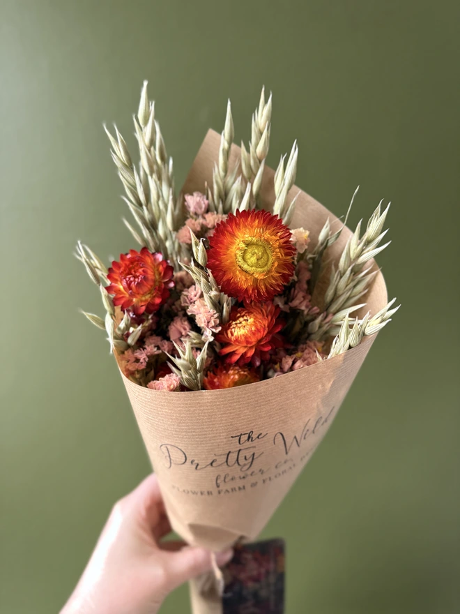 a bunch of dried flowers in bright colours wrapped in black tissue and kraft paper held by a person in a mustard coloured jumper with their left arm against a green wall